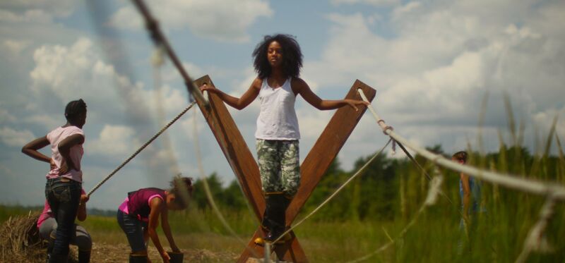 The image shows a group of people in a field. A woman stands on a wooden structure with her arms outstretched. Other people are working around her, some holding ropes. The sky is blue with some clouds.